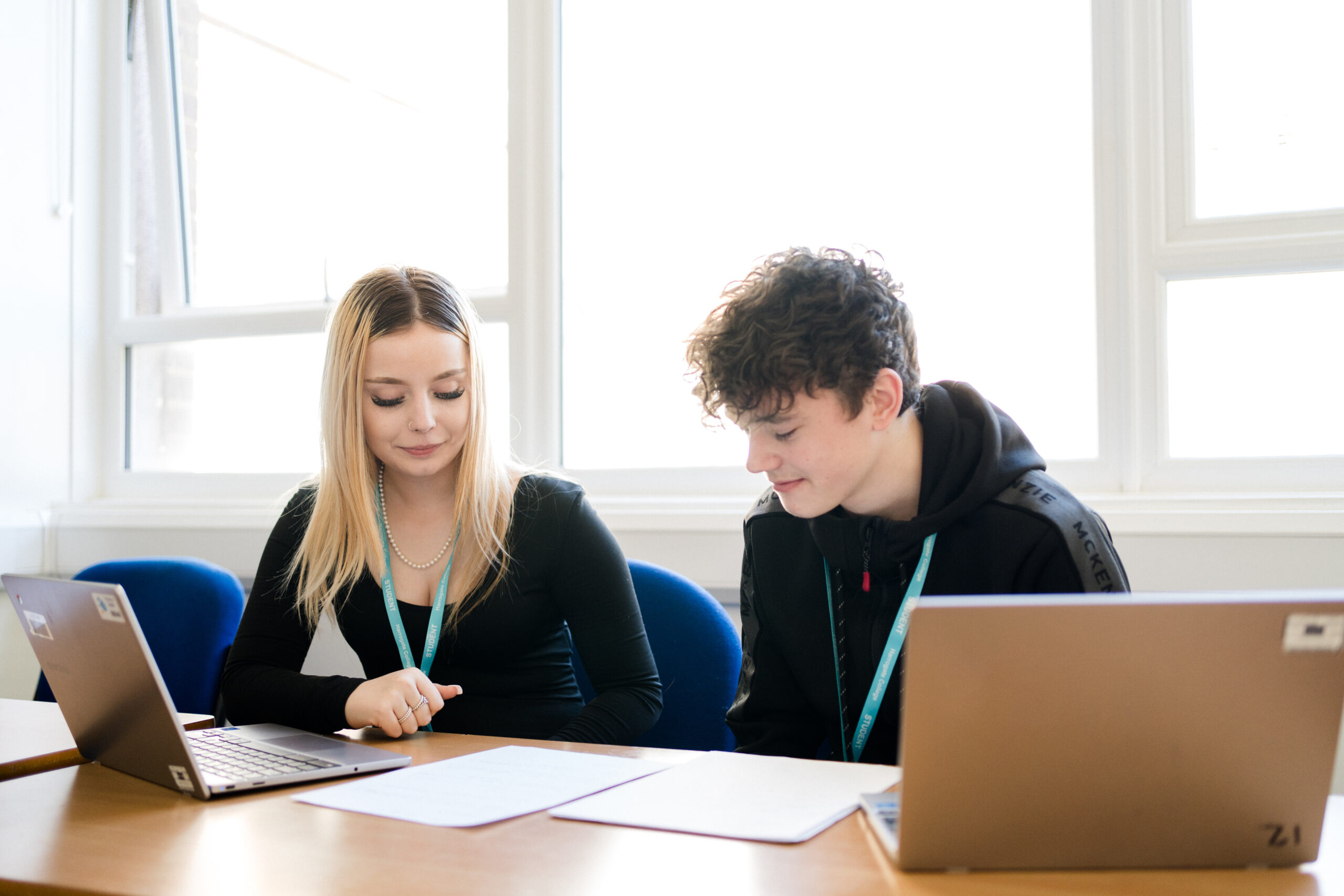 Two students wearing blue lanyard sitting in classroom looking at the notes with two laptops on the table