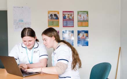 Two Harrogate College students wearing white college overalls sat at a desk in a classroom working on a laptop together smiling