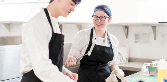 Two students wearing kitchen overalls and aprons in the training kitchens at Harrogate College