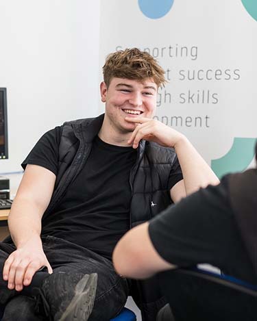 Student sat in Harrogate College smiling with hand on chin, in conversation with someone opposite